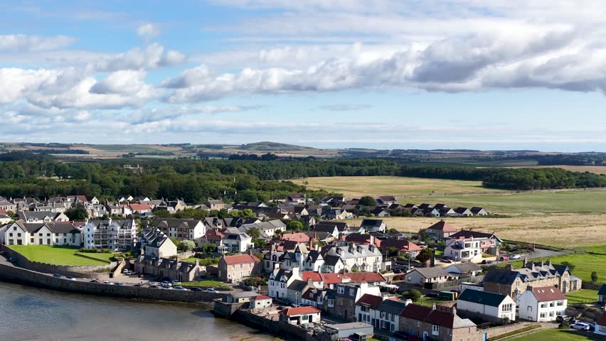 Drone pans above residential houses, countryside, and shoreline under bright daylight in Dundee, Scotland
