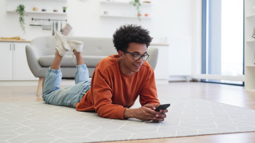 A young man with glasses lies on the floor, smiling while using his smartphone in a cozy living room.