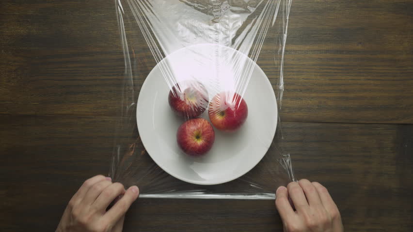 Woman hands are wrapping fresh ripe apples in cling film. Red fruits on white plate wrapped in plastic wrap. Food freshness concept. Protection for food.