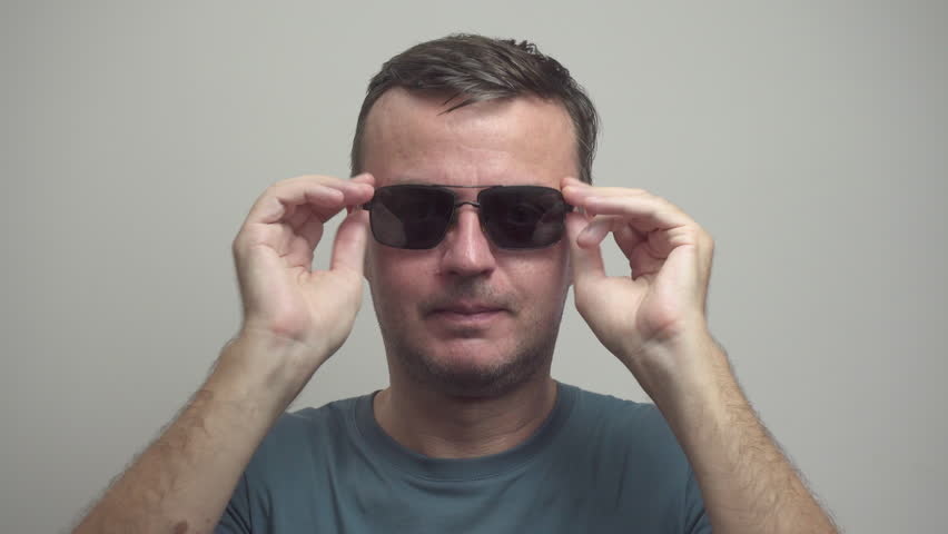 Middle-aged man is putting on sunglasses. Portrait of satisfied person in sunglasses on white wall background.