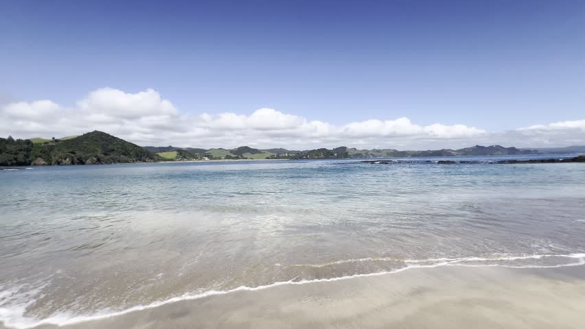 Clear waves roll onto the sandy shore at Whale Bay, New Zealand, with turquoise water, green hills and soft clouds under a bright blue sky. Peaceful coastal scenery perfect for travel content.