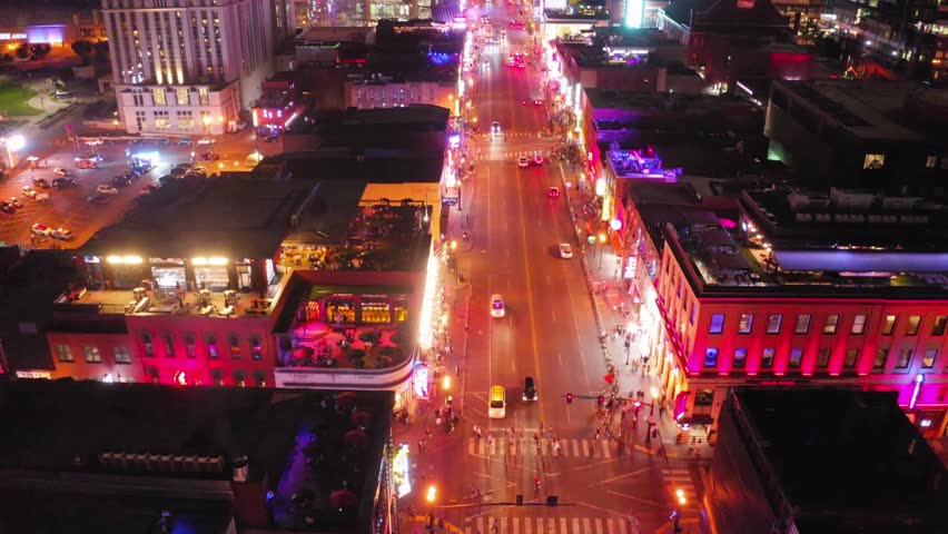 A high aerial view tilts up over Nashville's famous Broadway at night. Vibrant neon lights illuminate the street, busy rooftop bars, and city skyline.