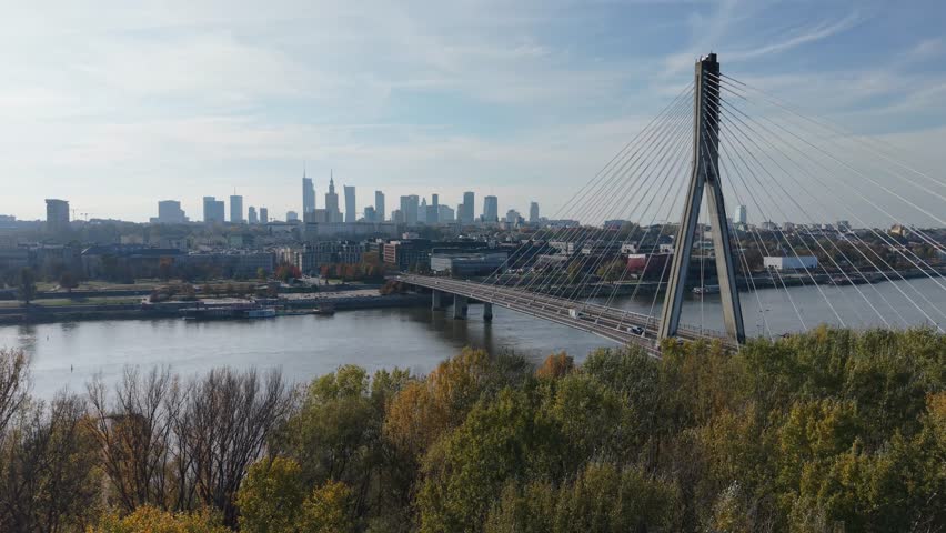 Aerial drone flyby near the Vistula River and its bridge in Warsaw, Poland, capturing the city skyline and reflections on the calm water.