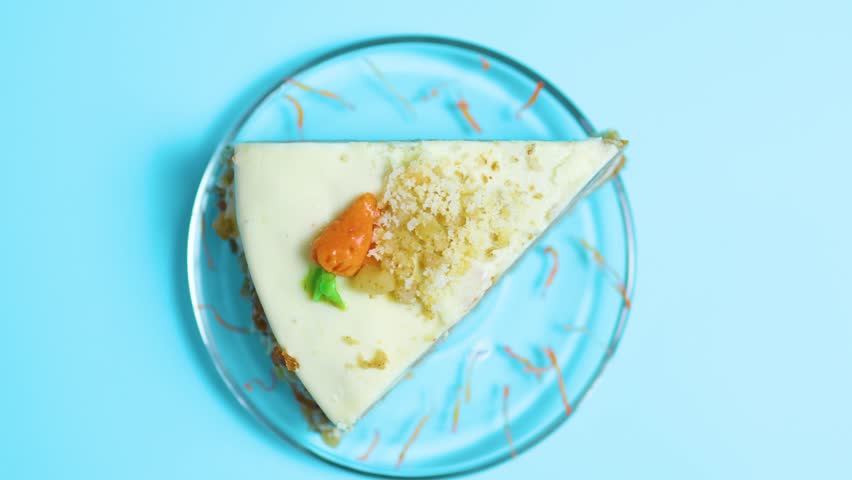 Carrot cake slice on a clear glass plate against a vibrant blue background, topped with cream cheese frosting, crunchy nuts, and a small decorative carrot, presenting a sweet dessert