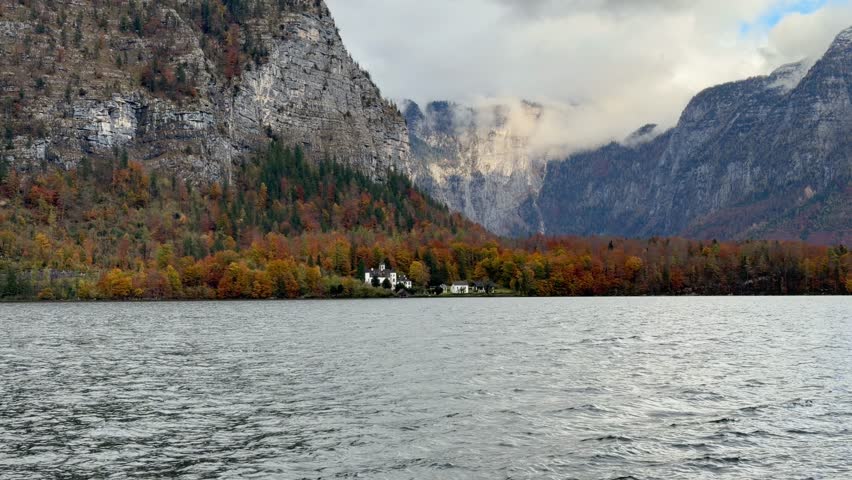 Panoramic view of Lake Hallstatt and the dramatic Austrian Alps in autumn. A solitary white house is nestled among the colorful fall trees at the foot of steep, rocky cliffs.