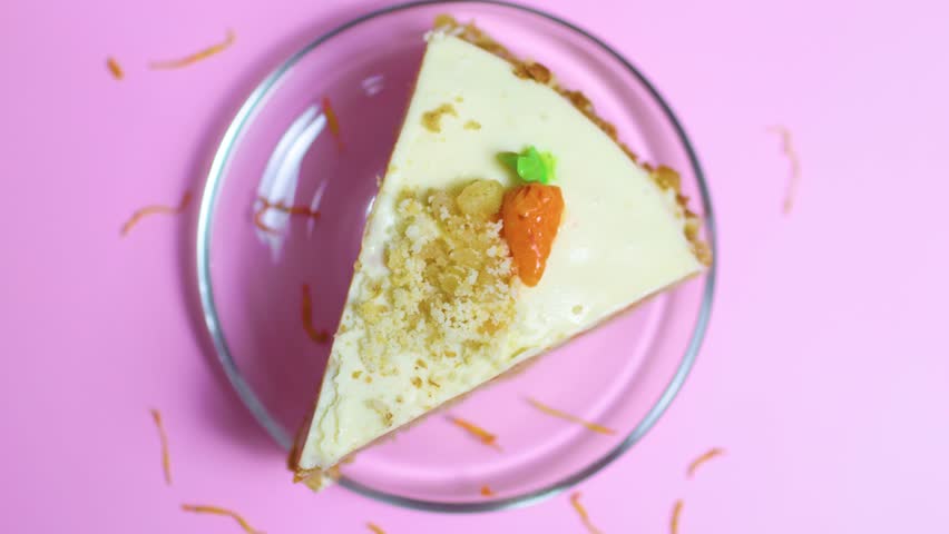 Slice of classic carrot cake with rich cream cheese frosting. Topped with crushed nuts and a small marzipan carrot. Resting on a glass plate against a bold pink background. Representing a sweet treat