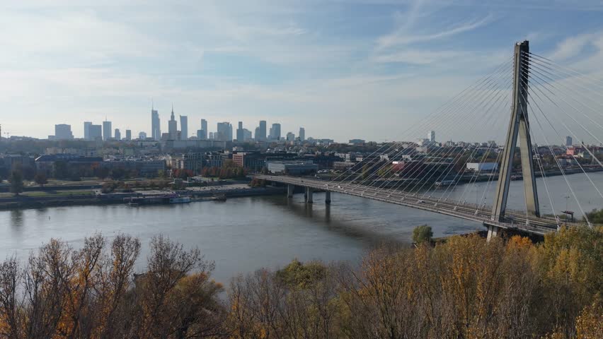 Aerial drone footage crossing the Vistula River near the bridge in Warsaw, Poland, revealing the modern skyline and reflections of the city in the water.