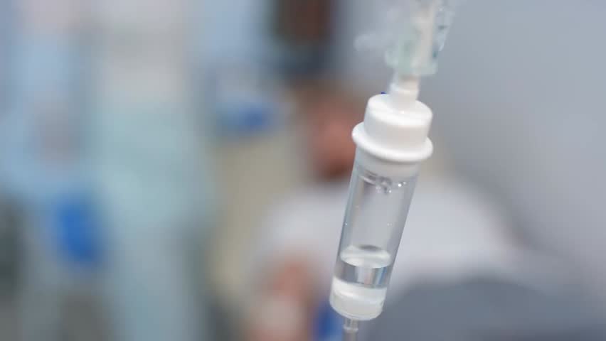 Selective focus on IV drip in hospital during intravenous therapy, with nurse attending a male patient in the background, highlighting medical care.