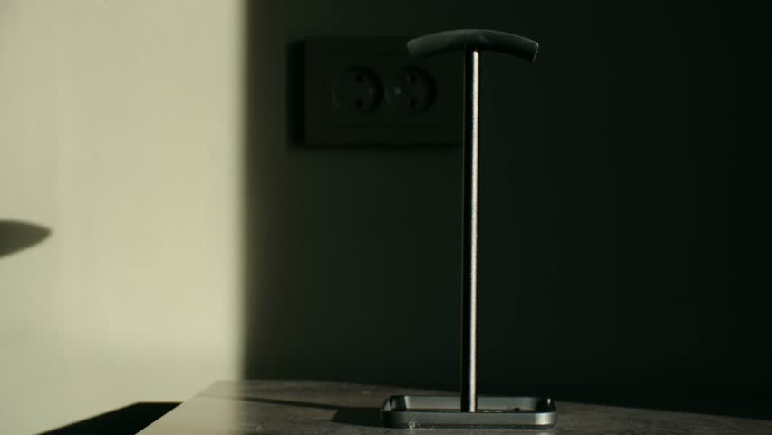 Person hand placing a pair of modern black wireless headphones on a special stand located on a desk, with dramatic sunlight creating strong shadows and highlights in the background