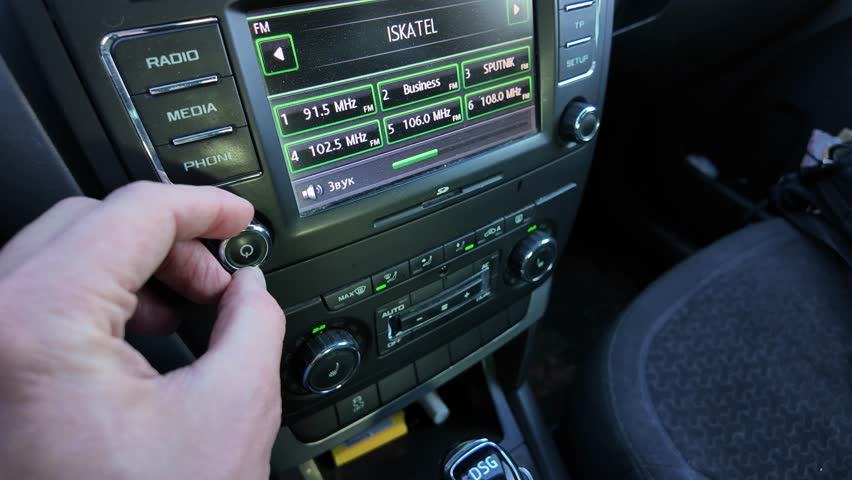 hand adjusting car radio knob on dashboard, closeup of console display showing radio presets, finger rotating volume dial, illuminated buttons and textured knob, calm morning drive atmosphere