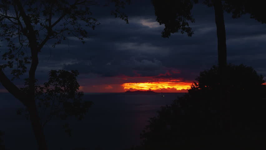 Woman Walks Toward the Edge of a Cliff Surrounded by Trees as She Watches the Fiery Sunset Over the Ocean in Silhouette Framed by Dark Clouds in a Wide Angle Scene.