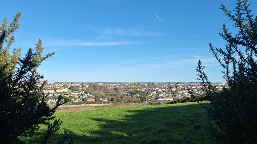 A calm hilltop pan moves across a flowering gorse bush before revealing a broad view of distant houses, open fields and a clear blue sky with forming clouds, finishing back on the landscape below.