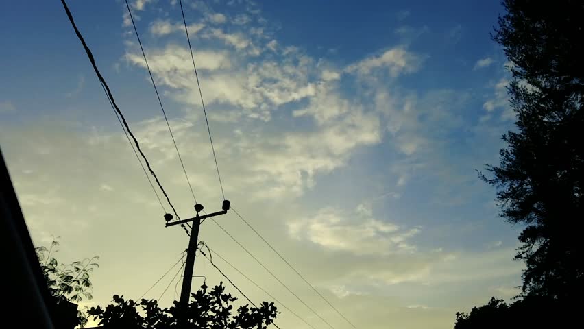 Blue sky with clouds, tree silhouettes, and electric power lines during sunset	