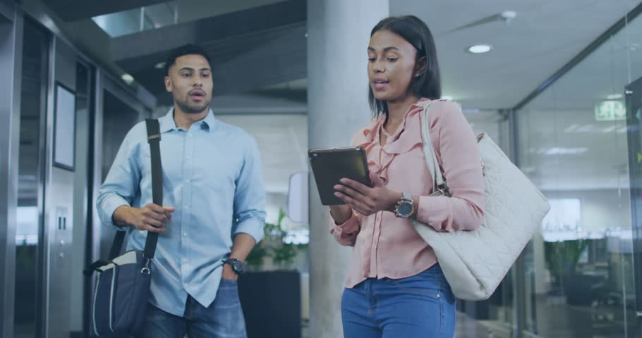Woman tapping tablet and activating AR overlay over screen, man leaning in reviewing business data. Lobby, glass, column, holographic, augmented, interactive, collaboration