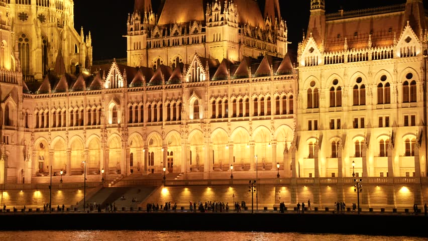 Detailed Close-Up footage of the Magnificent Hungarian Parliament Building in Budapest at Night, Featuring the Illuminated Gothic Revival Facade and Tourists on the Promenade.
