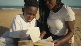 Caring african american mother sitting with son on sandy beach, teaching him how to read book together during sunny day, fostering education, family bonding, and parenthood - Powered by Shutterstock - Get 15% off with code: PIKWIZARD15