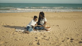 Loving african american mother and young son sitting on blanket on  sand, enjoy peaceful day by sea while read storybook together during their family picnic on sunny day - Powered by Shutterstock - Get 15% off with code: PIKWIZARD15