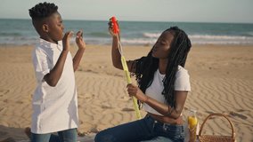 Young black woman with son enjoy summer day, blowing soap bubbles together during picnic on sandy beach. Happy family bonding and playing outdoors by sea during vacation - Powered by Shutterstock - Get 15% off with code: PIKWIZARD15