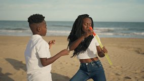 Joyful african american mother and young son having fun together on sandy beach, with woman blowing large soap bubbles and laughing boy trying to catch them during sunny day - Powered by Shutterstock - Get 15% off with code: PIKWIZARD15