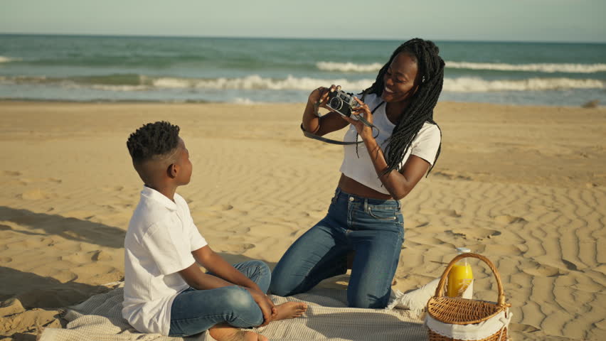 Young african american mother taking pictures of her son with vintage film camera during family picnic on sunny beach, enjoying summer vacation and creating memories together