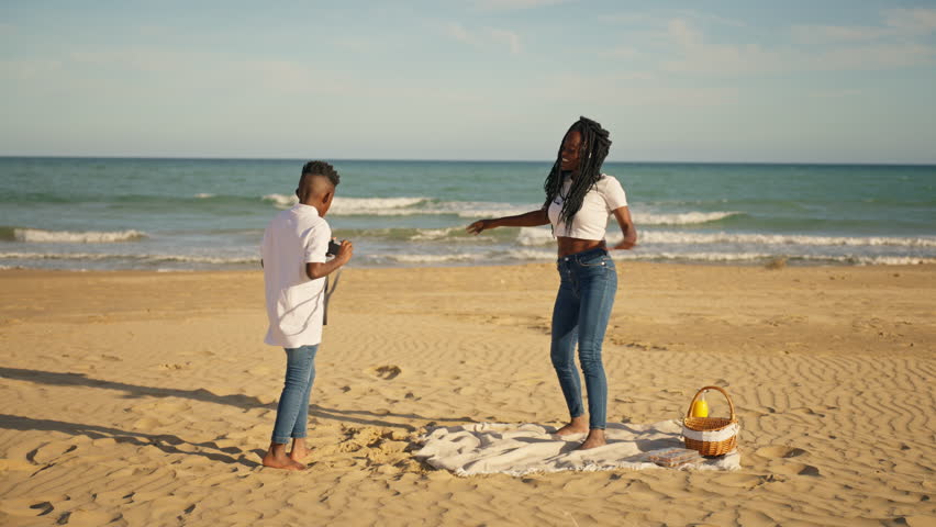 Joyful african american woman with stylish long braids striking pose for photo as young boy captures moment with an old-fashioned film camera at delightful beach picnic along  shore