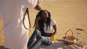 Young african american woman with long dreadlocks sitting on blanket and smiling while her boyfriend photographs during romantic picnic on sandy beach at sunset - Powered by Shutterstock - Get 15% off with code: PIKWIZARD15