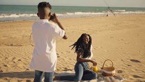 Beautiful young woman with dreadlocks smiling and posing for a photo while having a picnic on a sandy beach, as her younger brother or son takes her picture with an analog camera on a sunny day - Powered by Shutterstock - Get 15% off with code: PIKWIZARD15