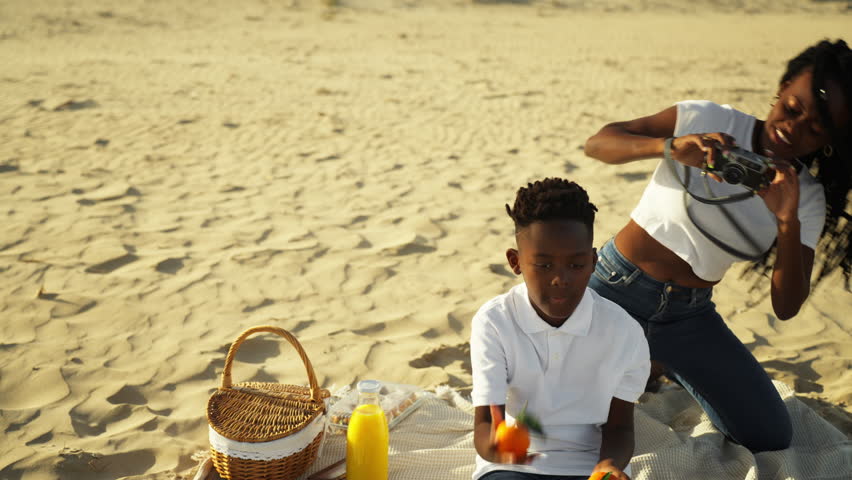 Joyful african american mother taking photos with vintage camera of son juggling oranges during sunny family picnic on sandy beach, capturing happy childhood moment outdoors