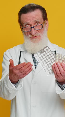 Senior doctor cardiologist man shows lot of tablet pills in blisters recommends drugs vitamin antibiotic cure treatment. Health care. Apothecary pharmacist grandfather isolated on yellow background