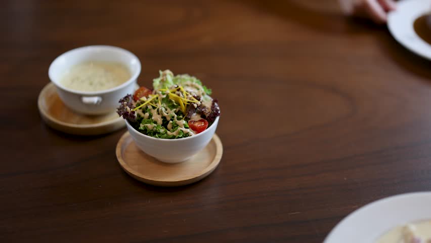Hand places salad next to omelette rice and creamy soup on wooden table in daylight.