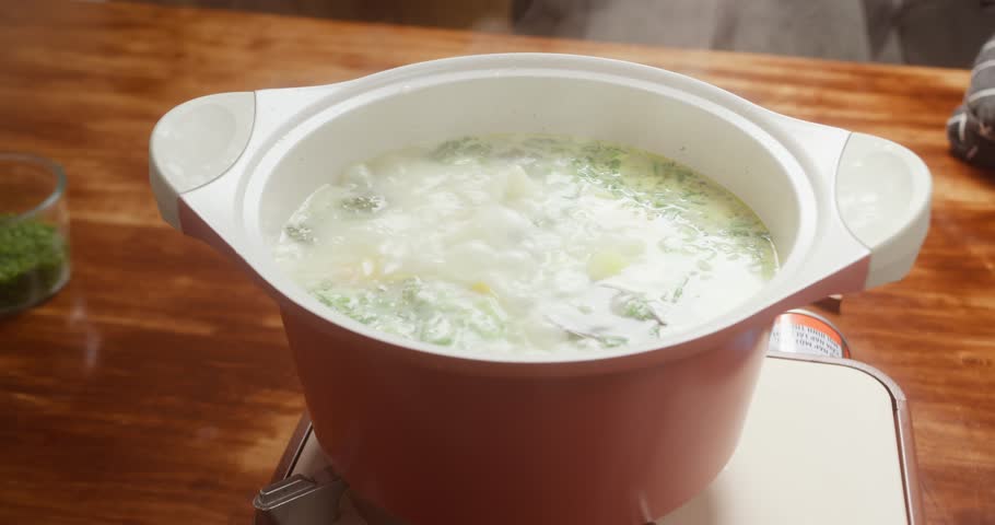 Woman cooking broccoli soup and adding chopped green onions, Female hands adding spring onions to boiling broccoli soup, Broccoli cream soup with vegetables boiling in pot.