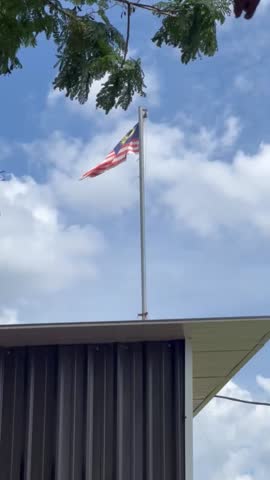 A low-angle video clip of the Malaysian flag, the Jalur Gemilang, billowing proudly in the wind from a flagpole mounted on the corrugated metal roof of a building.