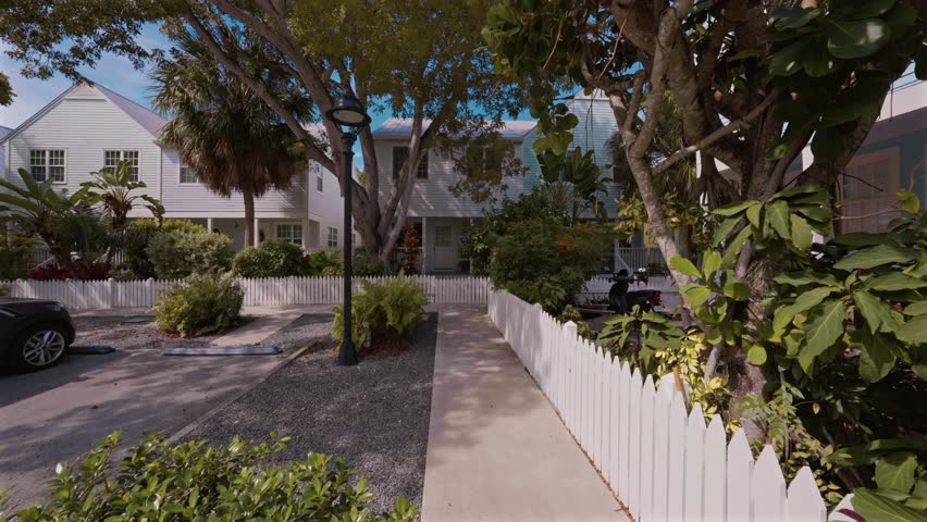 POV walk along a tropical residential path lined with white picket fences, lush greenery, and charming pastel homes in a peaceful coastal community