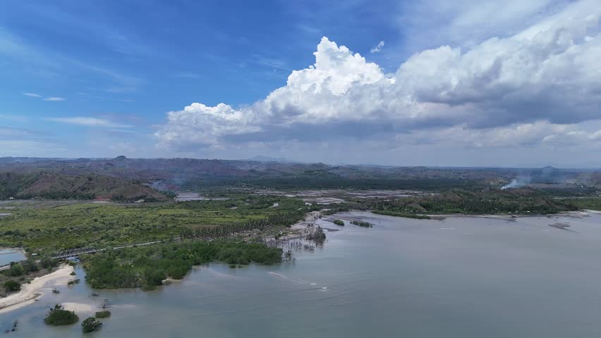 Aerial view of a vast tropical coastal landscape with lush green forests, hills, a wide bay, and dramatic cumulus clouds in a bright blue sky, showcasing natural beauty.
