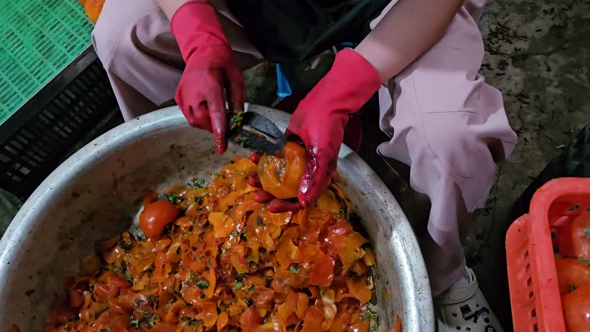 Close up of a woman wearing red protective gloves is peeling the skin of ripe persimmons over a large metal basin.	