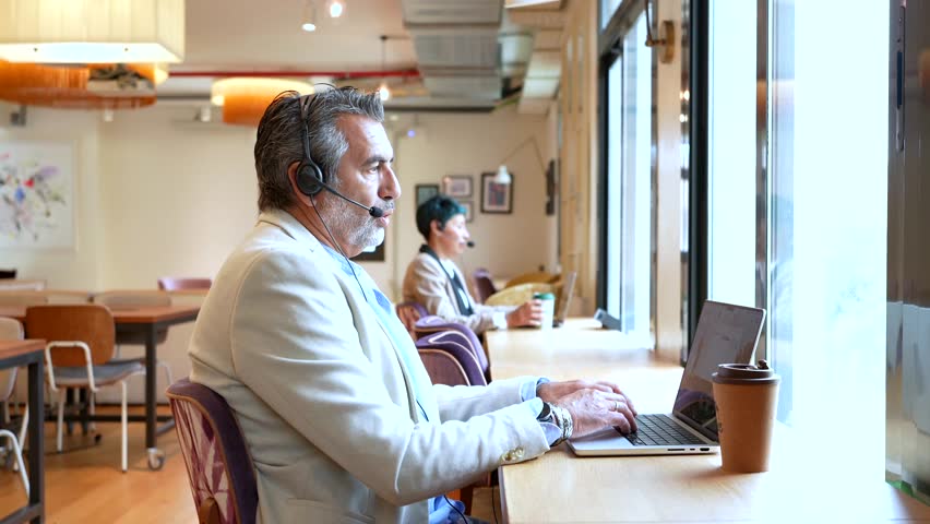 Focused mature businessman wearing a headset and talking during a video conference on his laptop, sitting at a desk in a modern coworking office space with a colleague in the background - Powered by Shutterstock - Get 15% off with code: PIKWIZARD15