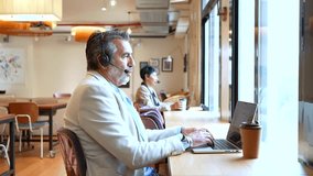 Focused mature businessman wearing a headset and talking during a video conference on his laptop, sitting at a desk in a modern coworking office space with a colleague in the background - Powered by Shutterstock - Get 15% off with code: PIKWIZARD15
