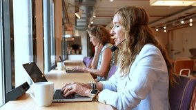 Businesswoman with a headset typing on a laptop and talking to a client while providing remote customer service and technical support in a modern, shared open plan coworking office space - Powered by Shutterstock - Get 15% off with code: PIKWIZARD15