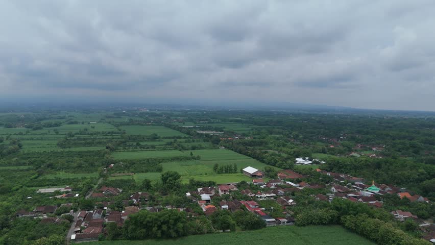 An expansive aerial panorama of a rural Indonesian landscape, featuring green farmland and forests bordered by scattered villages, with distant mountains under a dramatic, cloudy sky.