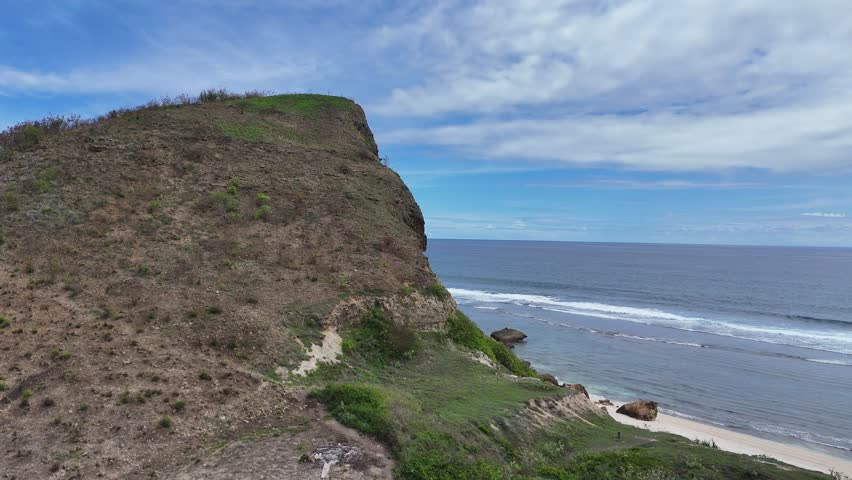 Dramatic Aerial View of Rugged Tropical Cliffside and Secluded Beach with Turquoise Ocean Waves Under a Bright Blue Sky on a Sunny Day