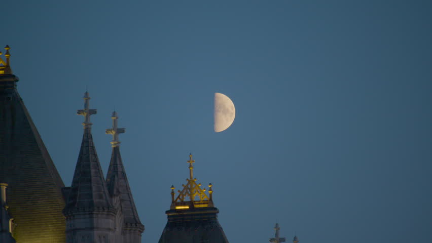 A bright half-moon hangs in a deep blue evening sky above the illuminated Gothic spires and golden-tipped pinnacles of Tower Bridge, creating a serene and dramatic night scene.