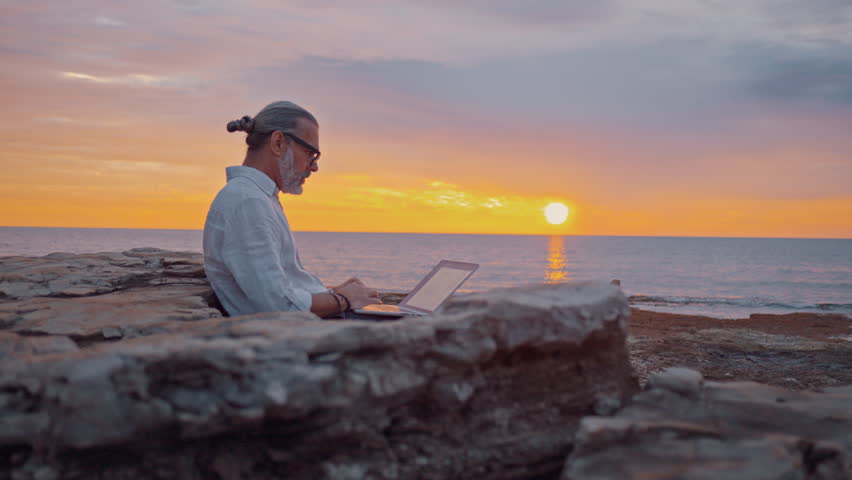 A focused digital nomad works on a laptop by the ocean at sunset, embodying remote work freedom. The serene coastal setting highlights productivity and tranquility. - Powered by Shutterstock - Get 15% off with code: PIKWIZARD15