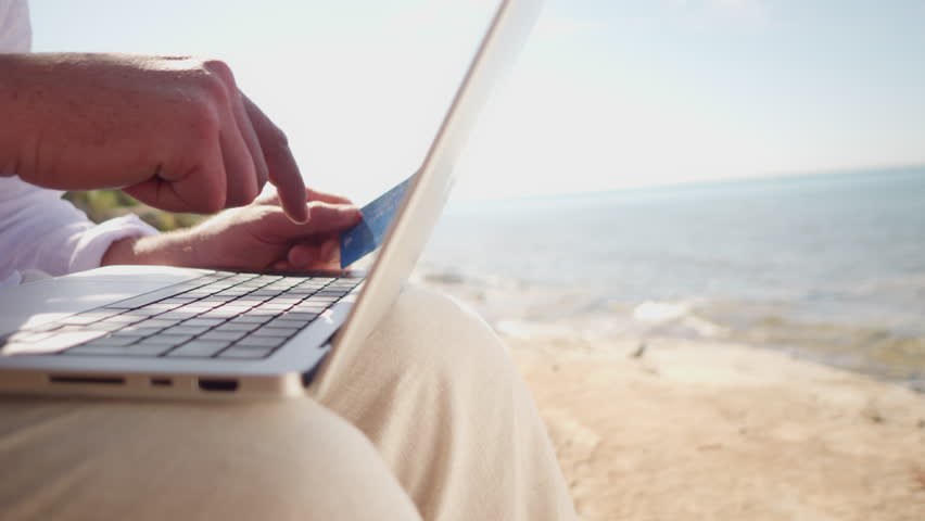 A digital nomad works on a laptop by the sea, holding a credit card, symbolizing remote work and financial transactions. The serene beach setting highlights the freedom and flexibility. - Powered by Shutterstock - Get 15% off with code: PIKWIZARD15