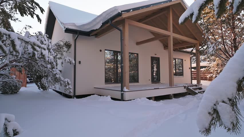 A modern house with a wooden porch stands in a snowy landscape. Snow-covered trees frame the house, creating a peaceful, winter scene