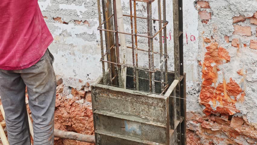 masonry worker pouring concrete mixture on shuttering box to create column at construction site. day time, stable shot, 4k.