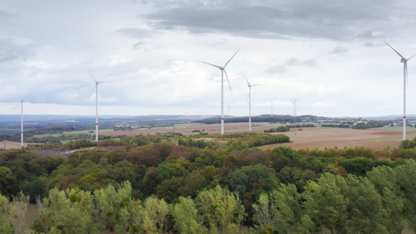 High Quality aerial view of wide fields with tall wind turbines above autumn trees calm sky renewable energy landscape in Luxembourg clean light soft colors and a peaceful cinematic atmosphere.