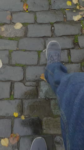 POV, person's legs going on damp granite pavers slabs covered with fallen yellow leaves. Top view of man whose feet are dressed in jeans and boots as he walks along wet sidewalk with autumn foliage