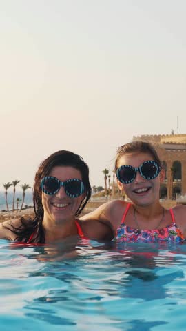 Woman and teen girl, mom and daughter, in funny, identical, round sunglasses, relax, have fun in outdoor pool with luxury hotel view. family vacation concept