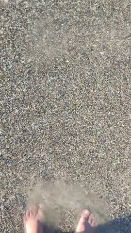A woman with bare feet standing on sand with shell on sandy beach on seashore. Waves of clean sea water rolling onto feet of white woman on sunny summer day. Top view. Rest, relaxation Vertical
