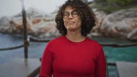 Woman with glasses and curly hair wearing red sweater closes eyes by stone edifice on wooden dock railing overlooking turquoise water; serenity calm reflection. - Powered by Shutterstock - Get 15% off with code: PIKWIZARD15